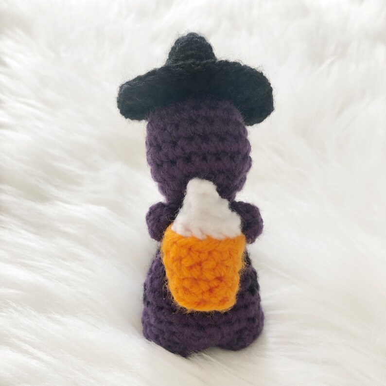 May include: A handmade crochet witch doll in purple, wearing a black hat, and holding a candy corn treat. The doll is crafted with visible stitches and set against a white, fluffy background, perfect for Halloween.
