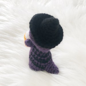 May include: A handmade crochet witch doll, featuring a black hat and a purple and black checkered dress. The doll is holding a small orange and white object, possibly a pumpkin or candy. The doll is set against a white, fluffy background.