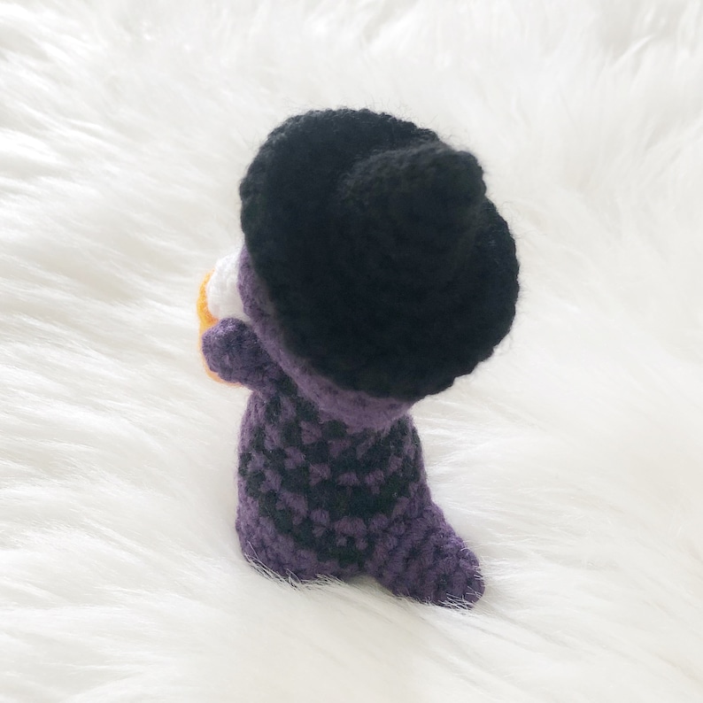 May include: A handmade crochet witch doll, featuring a black hat and a purple and black checkered dress. The doll is holding a small orange and white object, possibly a pumpkin or candy. The doll is set against a white, fluffy background.