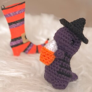 May include: A purple crocheted witch figure wearing a black hat, holding a candy corn bag. A crocheted orange and black striped witch boot is in the background. The Halloween-themed figures are set against a white, textured background.