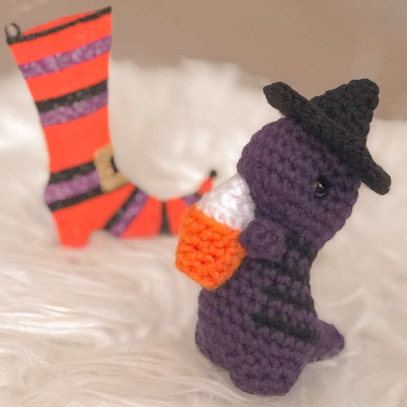 May include: A purple crocheted witch figure wearing a black hat, holding a candy corn bag. A crocheted orange and black striped witch boot is in the background. The Halloween-themed figures are set against a white, textured background.