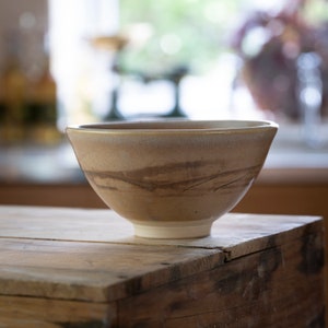May include: A large, light brown ceramic bowl with a white glaze and brown streaks. The bowl is sitting on a wooden surface.