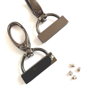 Könnte beinhalten: Zwei drehbare Karabinerhaken in Gunmetal-Farbe mit rechteckigen Platten und D-Ringen sowie vier kleine silberne Schrauben. Die Verschlüsse haben eine ovale Öffnung und einen federbelasteten Verschluss. Ideal für die Herstellung von Taschen und Bastelarbeiten.