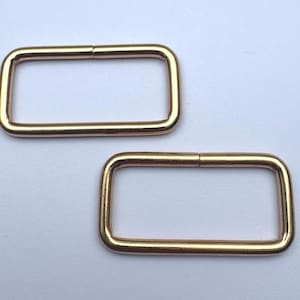 May include: Two gold-colored rectangular metal rings. Each ring has a rectangular shape with rounded corners and a small gap on one side. The rings are placed on a white surface.