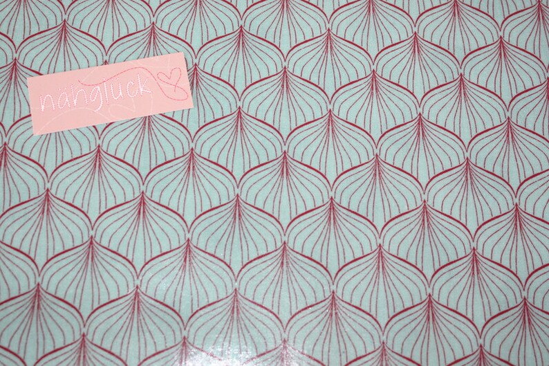 Puede incluir: Una tela azul claro con un patr&oacute;n repetitivo de formas de hojas granates. Una etiqueta rectangular rosa con la palabra "n&auml;hgl&uuml;ck" y un coraz&oacute;n est&aacute; colocada sobre la tela.