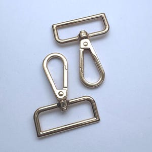 Könnte beinhalten: Zwei goldfarbene Karabinerverschlüsse aus Metall mit rechteckigen Schlaufen. Jeder Verschluss verfügt über einen federbelasteten Mechanismus und ein drehbares Gelenk, das den Haken mit der rechteckigen Basis verbindet und einen Befestigungspunkt bietet.