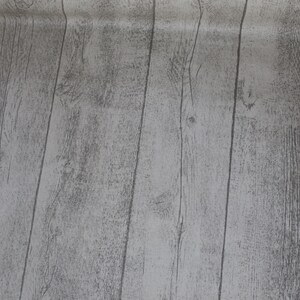 Könnte beinhalten: Eine Rolle hellgrauer Tapete in Holzoptik. Die Tapete zeigt vertikale Bretter mit sichtbarer Holzmaserung und Knoten. Die Farbe ist ein gedämpftes Grau mit dunkleren Grautönen, wodurch ein realistisches Holzbild entsteht.