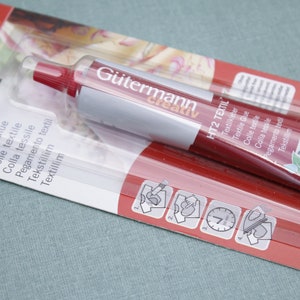 Könnte beinhalten: Eine rote und silberne Tube Gütermann HT2 Textil Stoffkleber. Die Tube ist in Deutsch und Englisch beschriftet. Die Verpackung zeigt ein weißes T-Shirt mit Anweisungen zur Verwendung des Klebers.