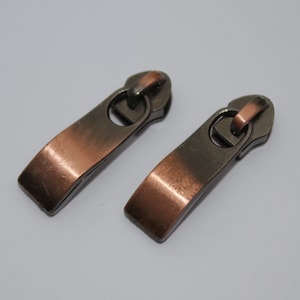 Könnte beinhalten: Zwei Reißverschluss-Zipper mit einer Kupfer- und Bronze-Oberfläche. Die Zipper sind rechteckig mit abgerundeter Oberseite und einer kleinen Schlaufe oben.