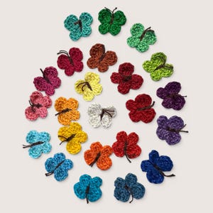 Op de afbeelding: Een verzameling van tweeëntwintig kleine, gehaakte vlinderapplicaties in diverse levendige kleuren, waaronder rood, blauw, groen, geel, oranje, paars, roze en wit. Elke vlinder heeft een donkerbruin gehaakt detail in het midden.