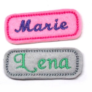 May include: Two felt name tags, one pink with purple embroidery that says "Marie" and one grey with green embroidery that says "Lena".
