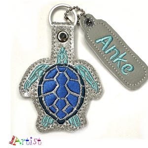 Könnte beinhalten: Ein silberner Glitzer-Schildkröten-Schlüsselanhänger mit einer blauen Schale und türkisfarbenen Akzenten. Der Schlüsselanhänger hat einen silbernen Ring und ein graues Leder-Etikett mit dem Namen "Anke" in Türkis gestickt.