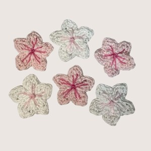 May include: Six individual five-petaled flowers crocheted from yarn in shades of white, pale pink, and deep magenta. Each flower features a central point where the yarn colors converge, creating a textured, layered appearance for these floral appliques.