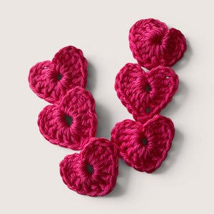 Könnte beinhalten: Sechs einzelne herzförmige Häkelapplikationen in leuchtendem Fuchsia-Pink. Jedes Stück ist mit einer sichtbaren Garnstruktur gefertigt und weist eine kleine offene Mitte auf, angeordnet in einer lockeren Gruppe vor einem hellen Hintergrund.