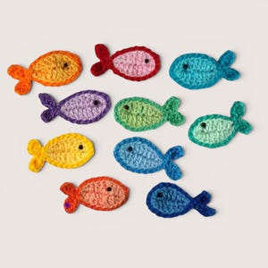 May include: A collection of ten small, flat, crocheted fish appliques in various colors including orange, red, pink, teal, purple, light green, yellow, blue, and dark blue. Each fish features a single small black bead for an eye.