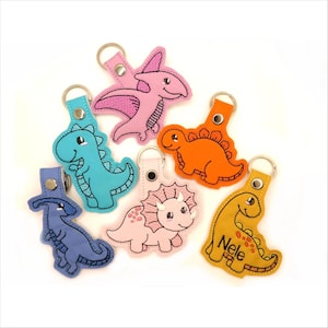 Può includere: Una collezione di sei portachiavi a forma di dinosauro in vari colori, tra cui rosa, blu, arancione, rosa chiaro e giallo. Ogni portachiavi è dotato di un anello in metallo e un bottone a pressione. Un portachiavi giallo riporta il nome Nele.