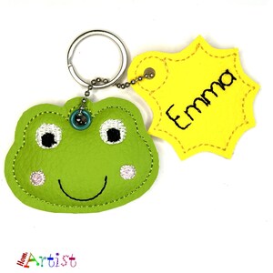 Può includere: Un portachiavi a forma di rana verde con un'etichetta gialla con scritto "Emma". La rana ha occhi bianchi, guance rosa e una bocca sorridente. Il portachiavi ha un anello e una catena argentati.
