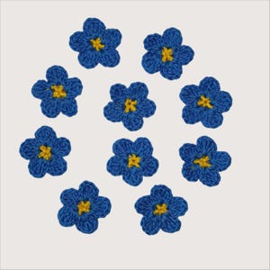 Puede incluir: Un conjunto de nueve flores individuales de cinco pétalos hechas de hilo azul con un pequeño centro amarillo. Cada flor está tejida a ganchillo con un patrón de puntos texturizado, dispuestas en formación circular sobre un fondo de color claro.