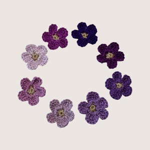 May include: A collection of seven individual crochet flower appliques arranged in a circular pattern. Each flower features five rounded petals in varying shades of purple, ranging from light lavender to deep violet, with a small tan center.