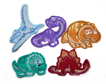 May include: Six felt dinosaur patches in various colors, including blue, purple, orange, green, and red. The patches are embroidered with details and have a whimsical, cartoon-like style.