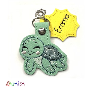 Può includere: Un portachiavi a forma di tartaruga turchese con un volto sorridente e un design a guscio grigio. Il portachiavi include un'etichetta gialla con il nome "Emma" in lettere nere, attaccata con una catena d'argento.
