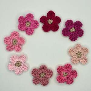 May include: A collection of eight individual crochet flowers arranged in a circle. Each flower features five rounded petals in varying shades of pink, ranging from light pastel to deep magenta, with a small beige center in each blossom.