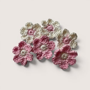 May include: A collection of six handmade crochet flower appliques. Three flowers feature white petals with a pink center, while the other three flowers feature pink petals with a white center. Each flower has five rounded petals and a textured yarn finish.
