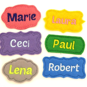 May include: Six colorful felt name tags with embroidered names: Marie, Laura, Ceci, Paul, Lena, and Robert. Each tag has a unique shape and color, including pink, yellow, purple, green, gray, and blue. The names are in contrasting colors.