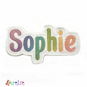 Puede incluir: Un parche bordado colorido con el nombre "Sophie" en colores arcoíris. El parche está hecho de fieltro y tiene un fondo blanco.