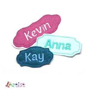 Puede incluir: Tres etiquetas de nombre de fieltro con nombres bordados. La primera etiqueta es rosa con el nombre "Kevin" en bordado blanco. La segunda etiqueta es azul claro con el nombre "Anna" en bordado turquesa. La tercera etiqueta es azul oscuro con el nombre "Kay" en bordado azul claro.