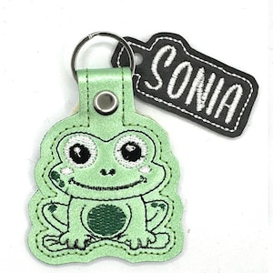 Peut inclure: Porte-clés en forme de grenouille verte avec une étiquette noire sur laquelle est brodé "SONIA" en blanc.