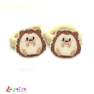 May include: Two cream-colored hair ties with embroidered brown hedgehogs. Each hedgehog has a white face with black eyes and a pink nose.