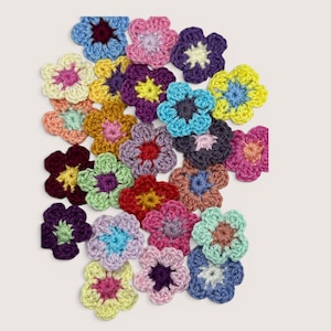 May include: A collection of twenty-four small, five-petaled crochet flowers in various colors including yellow, pink, blue, purple, red, green, and brown. Each flower features a contrasting center color, creating a vibrant and diverse assortment of handmade textile embellishments.