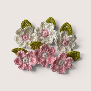 May include: A collection of six handmade crochet flower appliqués. Three flowers feature white petals with pink centers, and three feature pink petals with white centers. Each flower is accompanied by a small, textured green crochet leaf.