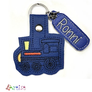 Puede incluir: Llavero de cuero azul con un diseño cosido de un tren y una etiqueta de cuero azul con el nombre "Ronni" cosido en blanco.