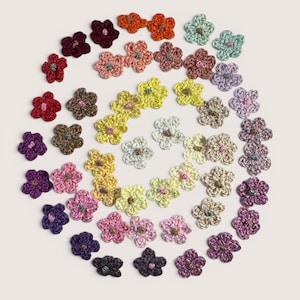 May include: A collection of small, five-petaled crochet flowers arranged in a spiral pattern. The flowers come in a variety of colors including red, orange, yellow, green, blue, purple, pink, white, and brown, each featuring a small center detail.