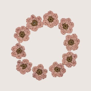 May include: A circular arrangement of nine individual crochet flowers. Each flower features five rounded petals in a soft dusty pink yarn with a contrasting dark olive green center. The crochet work displays a textured, looped stitch pattern.