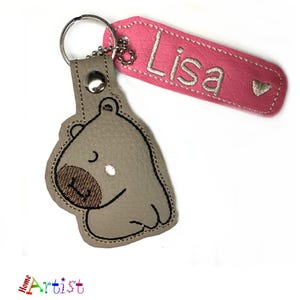 May include: A personalized keychain featuring a gray hippo design with a brown snout and a pink tag. The tag is embroidered with the name "Lisa" and a small heart. The keychain is attached to a silver key ring.