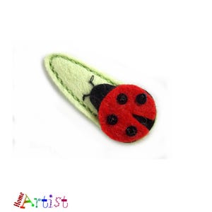 Peut inclure: Une coccinelle en feutre rouge et noir avec des points noirs sur une feuille en feutre vert. La coccinelle est une pince à cheveux.