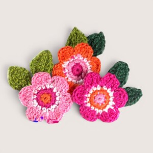 May include: Three crocheted flower appliques with five petals each, featuring vibrant color combinations of pink, orange, and white centers. Each flower is accompanied by textured green crocheted leaves, creating a layered floral arrangement.