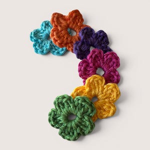 Könnte beinhalten: Eine Sammlung von sechs gehäkelten blumenförmigen Applikationen, die in einer geschwungenen Linie angeordnet sind. Die einzelnen Blumen sind in leuchtenden Farben wie Türkis, Orange, Violett, Magenta, Gelb und Grün gefertigt und haben eine zentrale Öffnung.