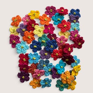 May include: A collection of small, five-petaled crochet flowers in a variety of vibrant colors including red, orange, yellow, blue, purple, and pink. Each individual flower features a small, bright lime green center accent stitched into the middle.