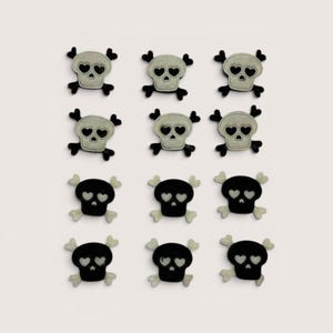 Könnte beinhalten: Ein Set aus zwölf gestickten Aufnähern, angeordnet in vier Reihen zu je drei Stück. Die Aufnäher zeigen Totenkopfmotive mit herzförmigen Augenhöhlen und gekreuzten Knochen. Sechs Aufnäher sind hellgrau mit schwarzen Details, sechs sind schwarz.