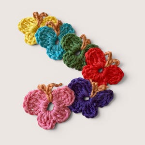 May include: A collection of six small, crocheted butterfly appliques arranged in a curved line. Each butterfly features a central body in a light tan yarn with wings crocheted in vibrant solid colors including yellow, teal, green, red, purple, and pink.