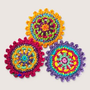May include: Three circular crochet coasters featuring intricate mandala-style patterns in vibrant colors including yellow, magenta, purple, turquoise, and lime green. Each coaster has a scalloped, gear-like edge and a complex, multi-layered design.