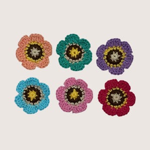 May include: A set of six crocheted flower appliques arranged in two rows. Each flower features five rounded petals in distinct colors including peach, teal, purple, light blue, pink, and dark red, with a dark brown and yellow center.