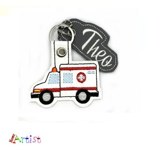 Può includere: Portachiavi ambulanza ricamato bianco e rosso con un'etichetta grigia che dice "Theo".