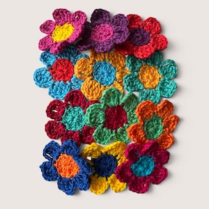 Könnte beinhalten: Eine Ansammlung von zwölf handgehäkelten Blumen in verschiedenen leuchtenden Farben wie Rot, Gelb, Blau, Lila, Orange und Grün. Jede Blume hat eine eigene mittlere Farbe und sechs Blütenblätter und ist in einer Gruppe auf einem schlichten Hintergrund angeordnet.