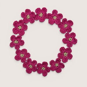 May include: A circular arrangement of twelve individual crocheted flowers. Each flower features five rounded petals in a vibrant magenta yarn with a small, contrasting light beige center. The flowers are connected to form a wreath-like shape.