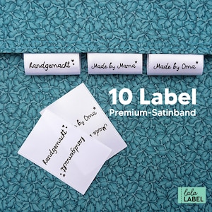 Könnte beinhalten: Zehn weiße Satinband-Etiketten mit schwarzem Text. Die Etiketten lauten "handgemacht", "Made by Mama", "Made by Oma" und "handgemacht by Oma". Die Etiketten befinden sich auf einem türkisfarbenen Hintergrund mit einem Blumenmuster.